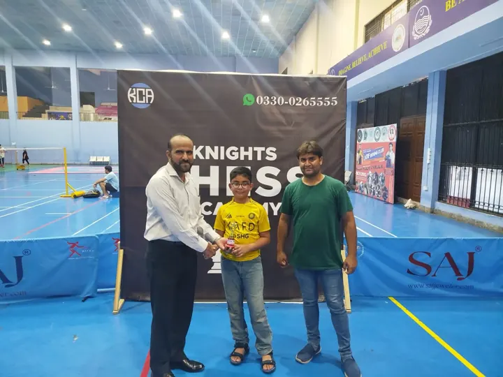 A boy in a yellow football shirt receives a small special-prize trophy between two organisers
