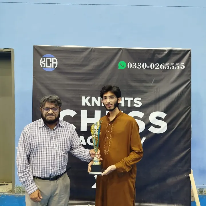 A young player in a brown kurta holds a gold trophy beside an organiser